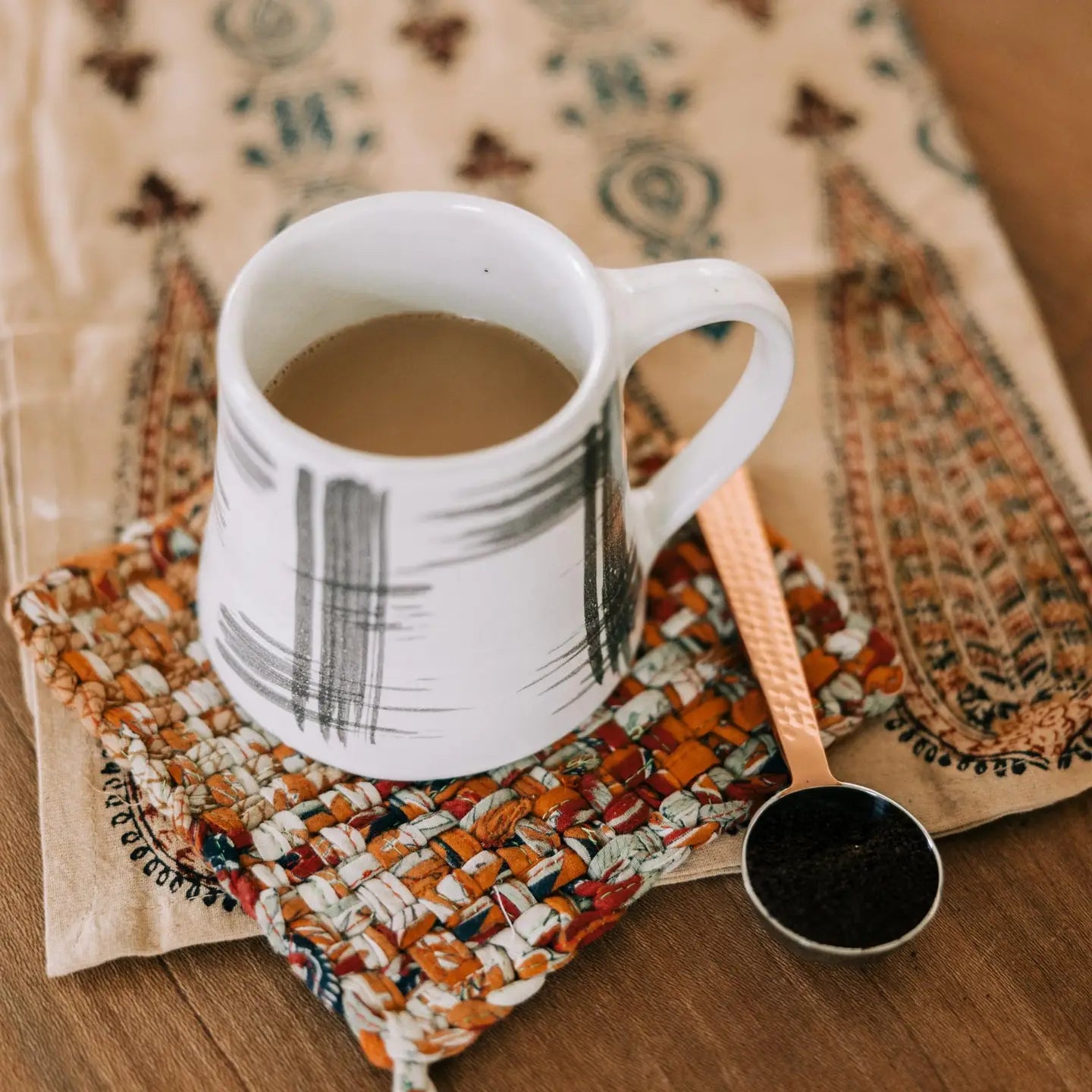 White mug with coffee on a patterned cloth with a hammered steel spoon nearby